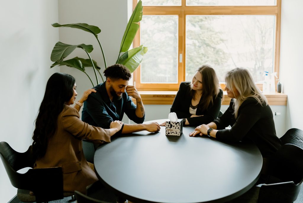 A picture of office workers sitting around table, one being upset, the rest being suppportive
