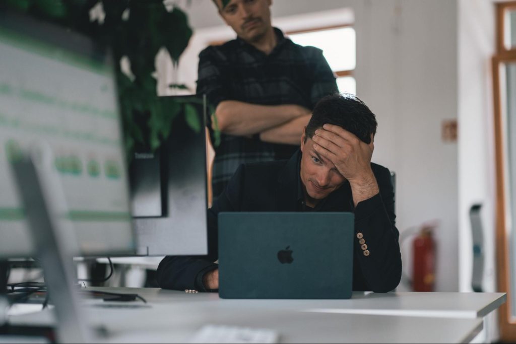 A man looking at laptop, head in hand, with other man looking over his shoulder