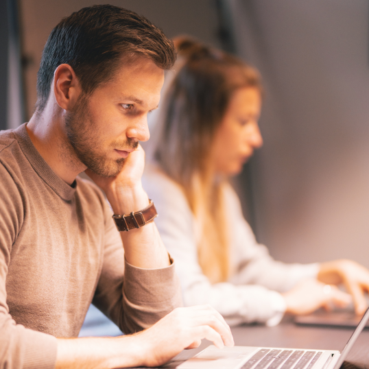 An image of two people sitting at a desk with a man in the foreground and a woman in the background.