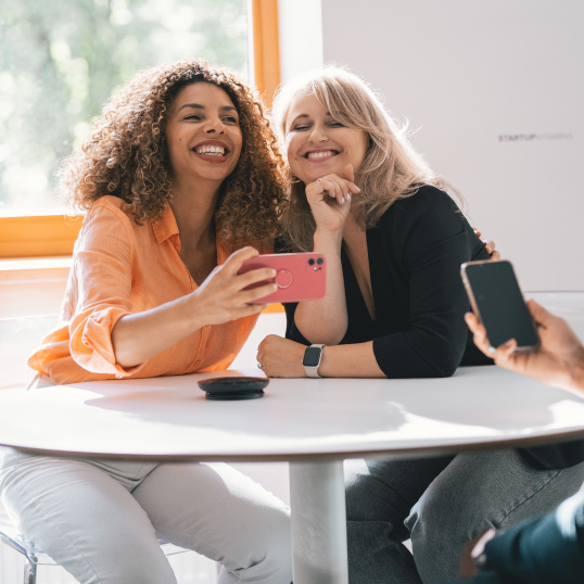 Imagen de dos mujeres sentadas a una mesa, inclinadas la una hacia la otra y sonriendo, con una de ellas sosteniendo un teléfono en la mano para hacer una foto.