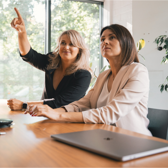 An image of two women sitting at a desk and one of the women is pointing towards something.