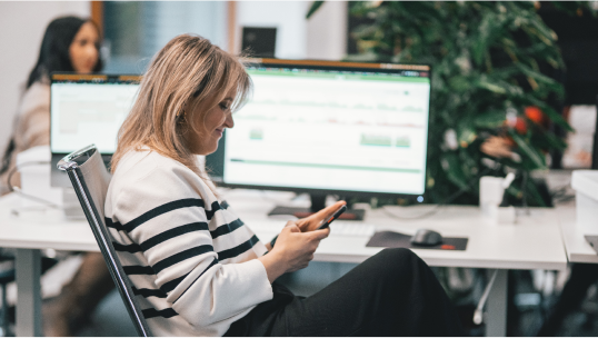 A woman using a phone in an office with a staff monitoring tool open on a computer screen in the background